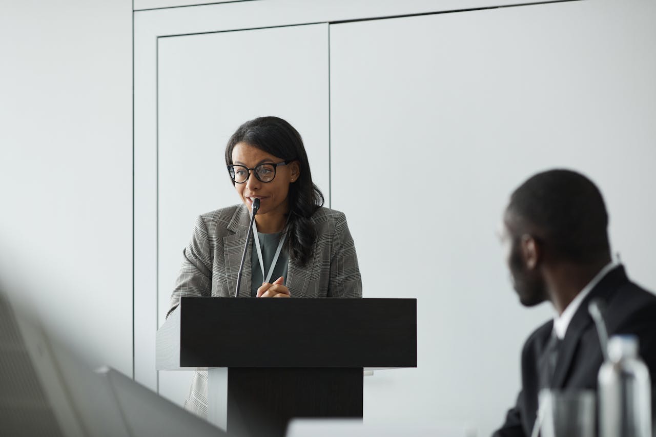 African American businesswoman delivering a speech at a conference podium with a colleague listening.
